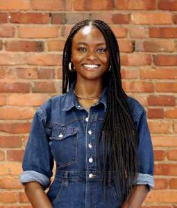 Samantha stands smiling in front of a brick wall, wearing a denim button shirt. 